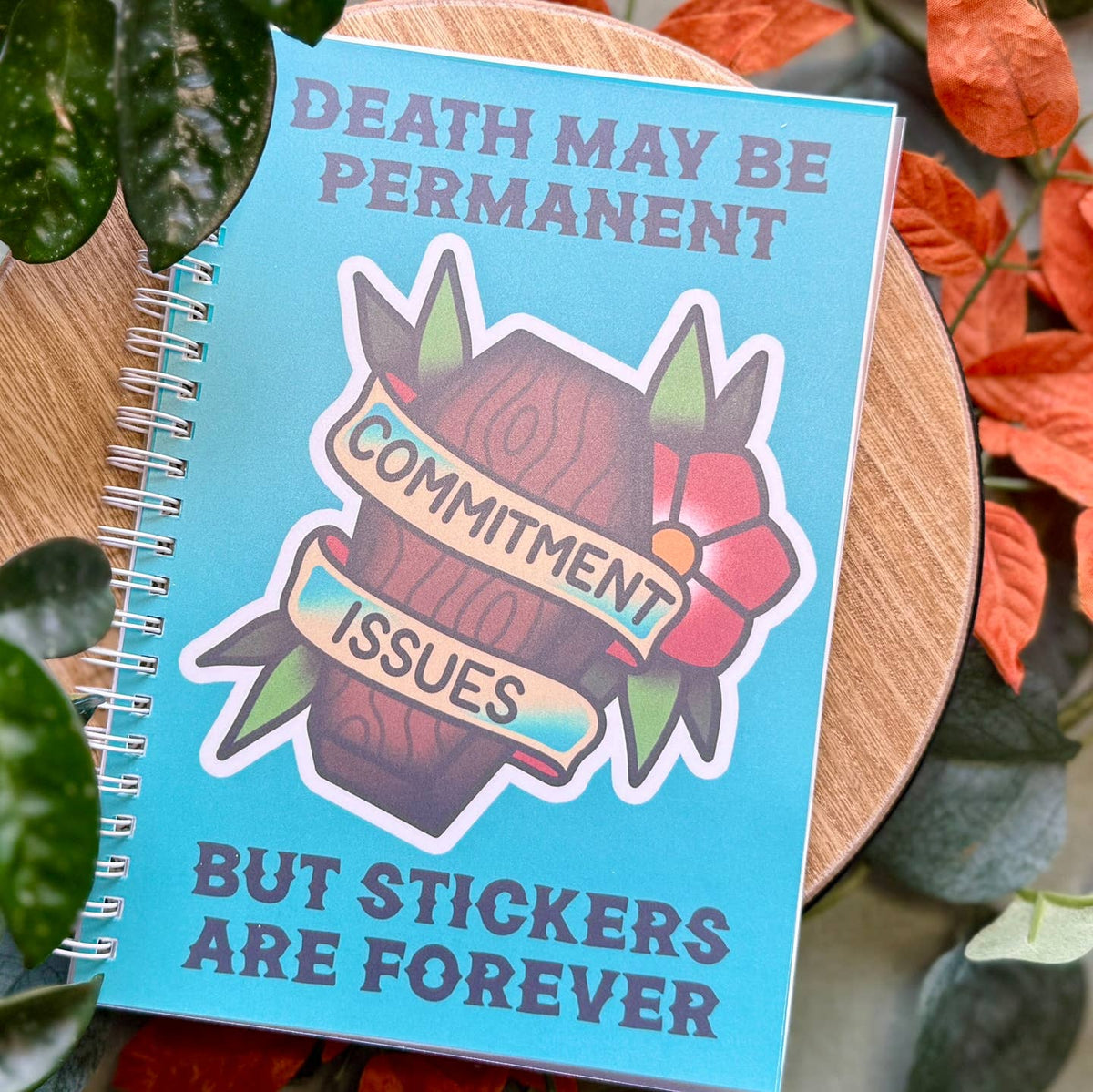 Spiral bound reusable sticker notebook with blue cover and tattoo illustration of a coffin, ribbon banner reading "Commitment Issues" and flower with text that reads, "Death May Be Permanent But Stickers Are Forever"