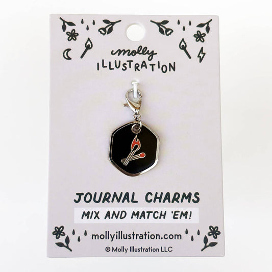 A black hexagonal journal charm with a silver clasp and a red-and-white match graphic, displayed on a white backing card labeled Molly Illustration.