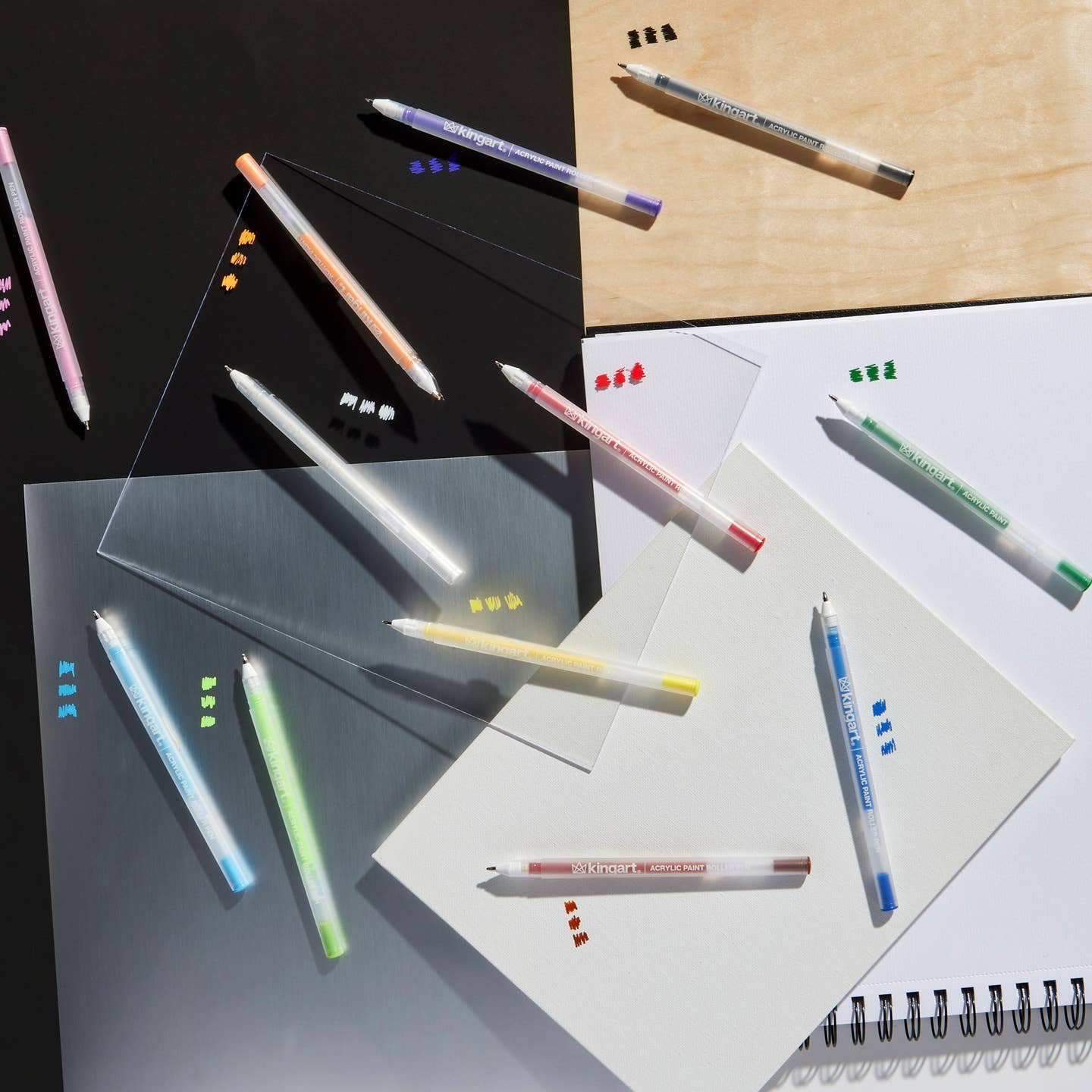 A styled flat-lay of the Kingart acrylic paint roller pens in 12 assorted colors, arranged diagonally showing the marks made on glass, acrylic. paper and wood.