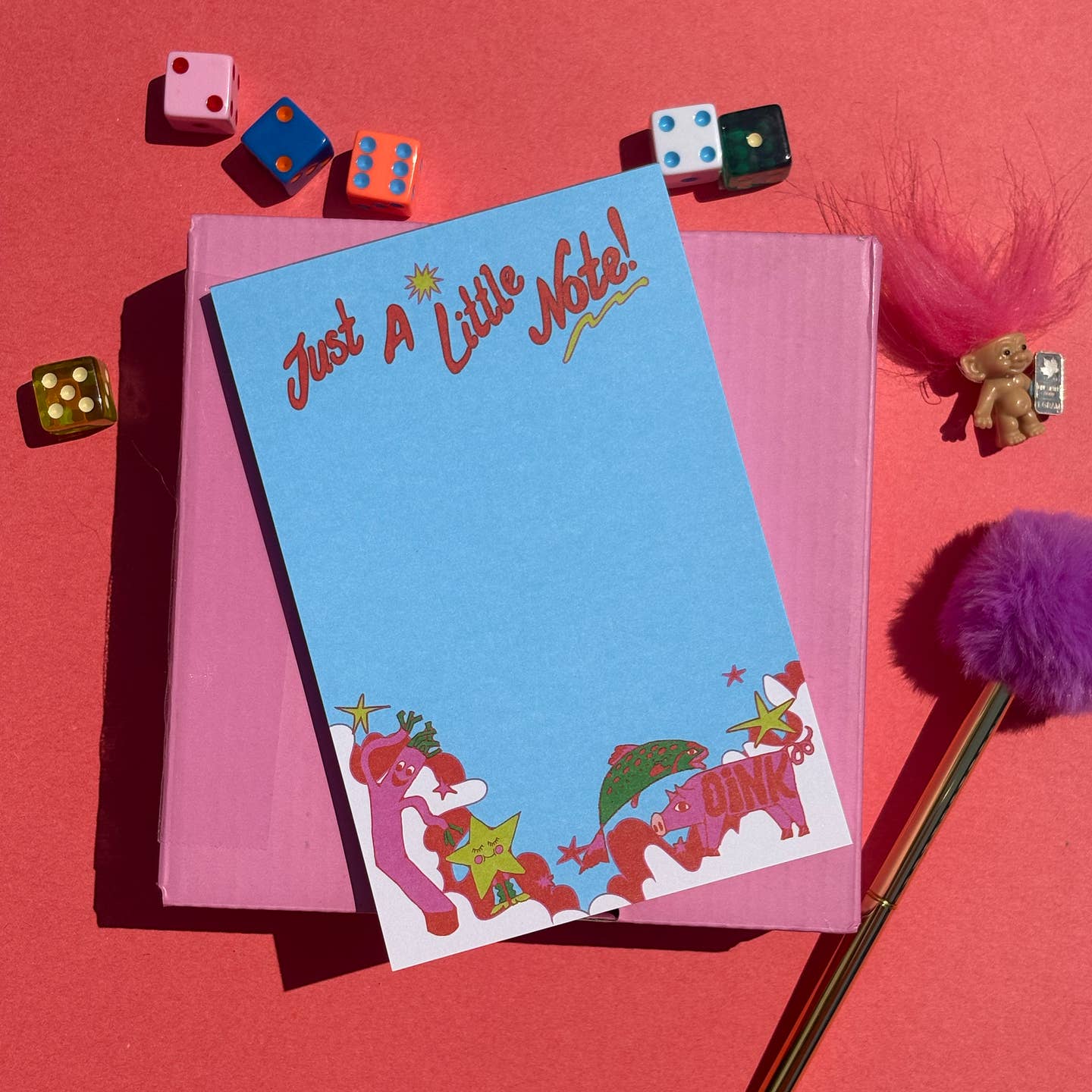 A flat-lay image of a pastel blue notepad with the text 'Just A Little Note!' and whimsical pink cartoon characters like a whacky waver and a pig at the bottom, on a red and pink background. The scene is styled with colorful dice, a purple pom-pom pen, and a small troll doll on a solid pink background.