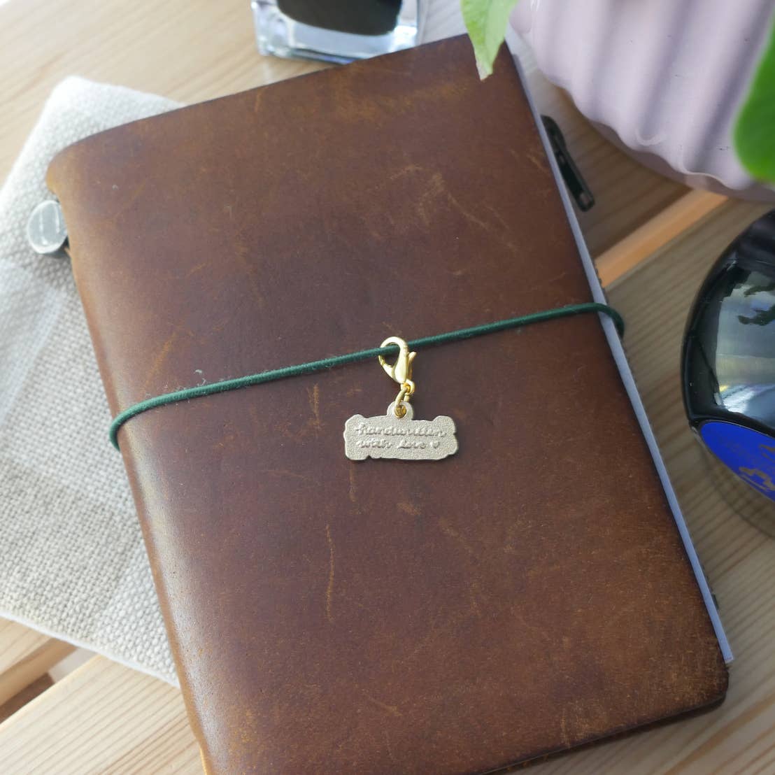 The back of the brass "Handwritten with Love" charm engraved with the words "handwritten with love" in a cursive style with a gold-tone clasp attached to a brown leather Traveler's style jounral.