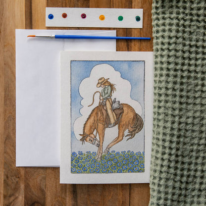 Card with a cowboy on a horse illustration, paintbrush, and color swatch on a wooden surface.