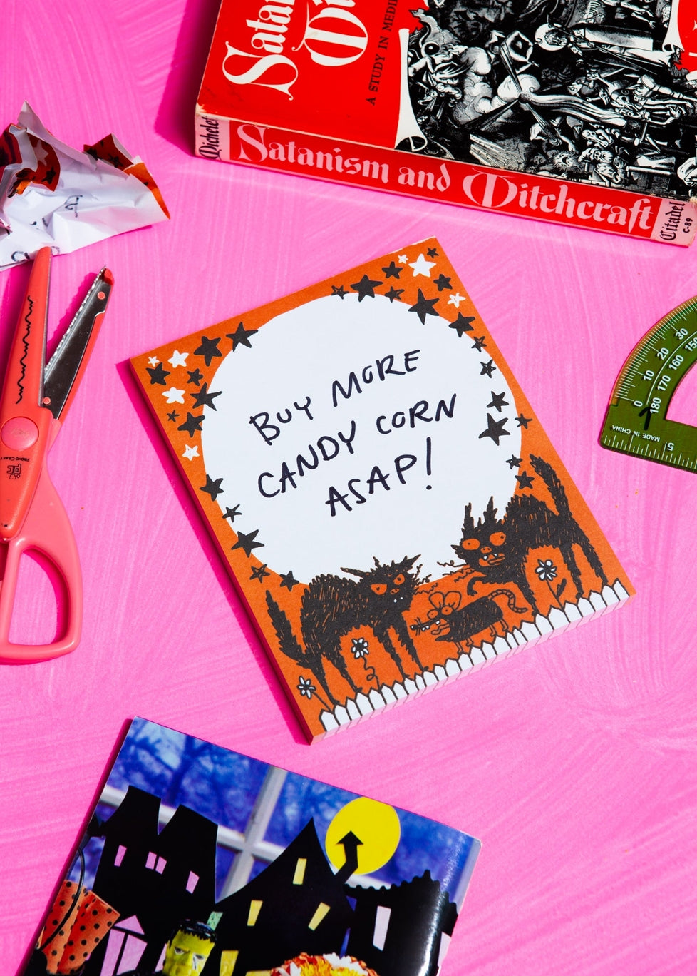 Large white moon makes up the body of this notepad with a deep orange border with slightly unhinged illustrations of two black cats and a rat. Someone has written "buy more candy corn asap!" on black marker on the notepad