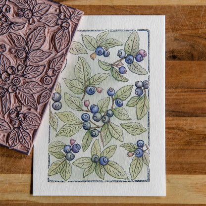 image of a lino block and a completed watercolor image of blue huckleberries and green leaves
