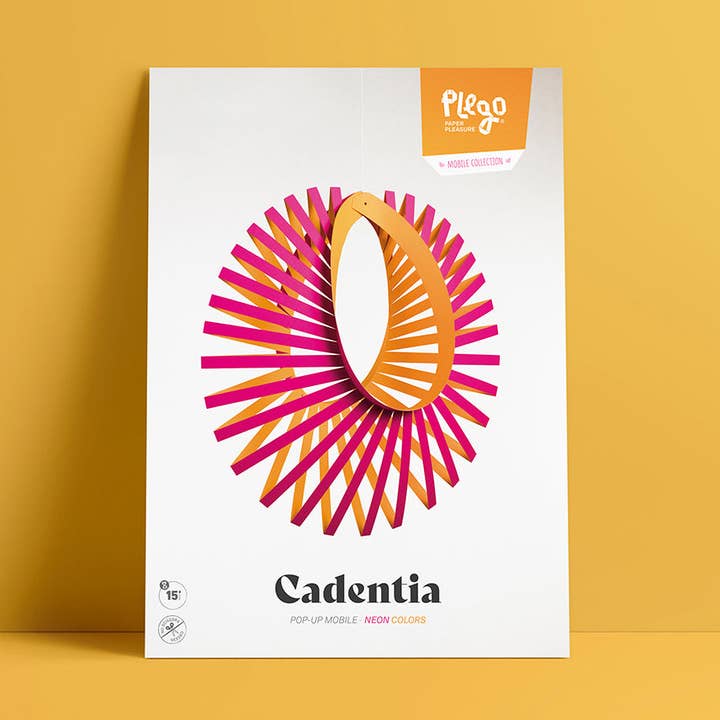 Cadentia Pop-Up Mobile by Plego, featuring a white rectangular box with a colorful geometric mobile in pink, orange, and yellow, set against a solid mustard yellow background.