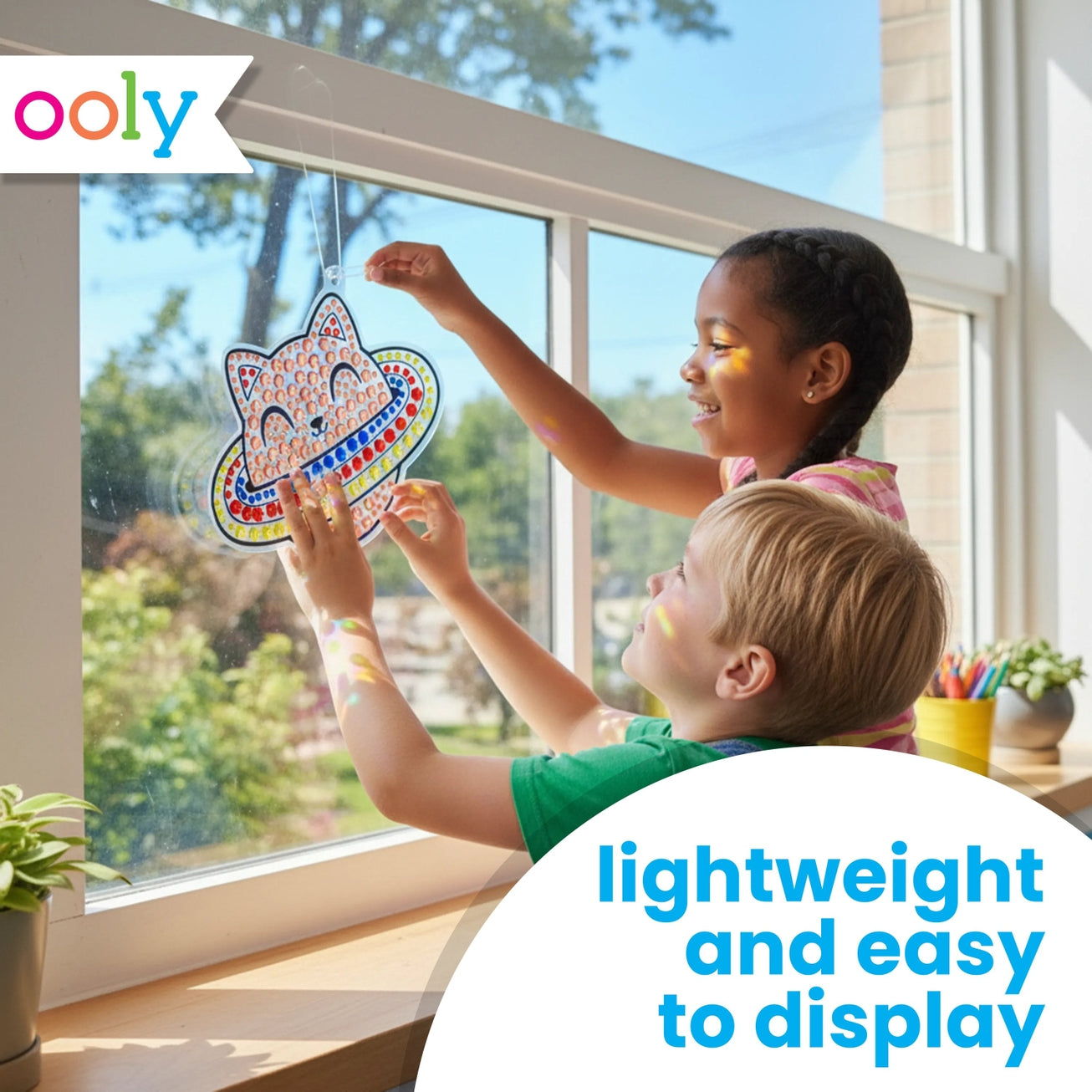 two children, one with white skin and short blond hair and one with brown skin and brown hair in two braids, hanging up a cat in space suncatcher in a brightly illuminated window. On a white background, blue text reads, "lightweight and easy to display"