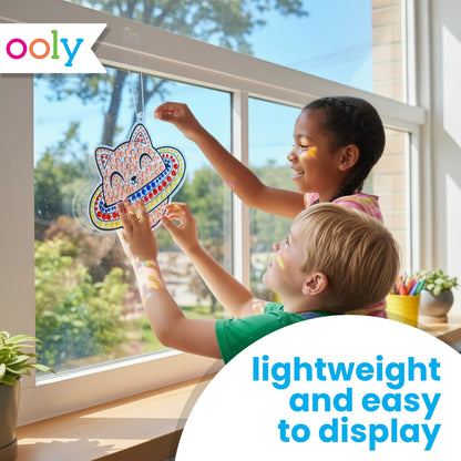 two children, one with white skin and short blond hair and one with brown skin and brown hair in two braids, hanging up a cat in space suncatcher in a brightly illuminated window. On a white background, blue text reads, "lightweight and easy to display"
