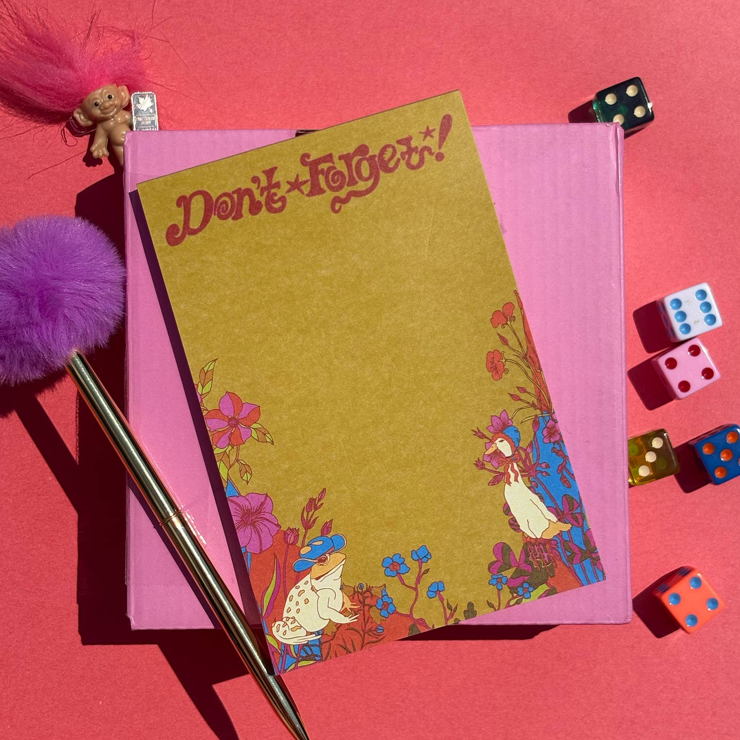A flat-lay image of a kraft color notepad with a whimsical floral border with a duck wearing a bonnet and a from wearing a cowboy hat, and the words 'Don't Forget!' in red script, placed on top of a pink notebook. A gold pen, purple pom-poms, dice, and a small troll doll are arranged around it on a pink background.