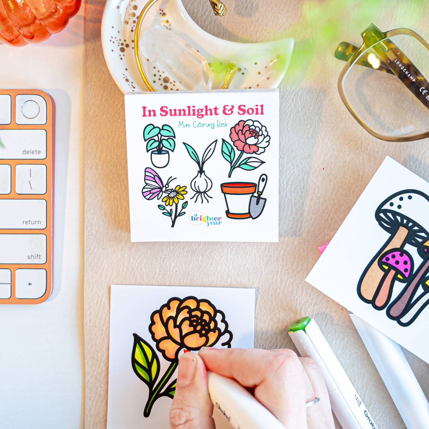 a hand in the process of coloring in a page featuring an pastel orange dahlia and pink and brown mushrooms