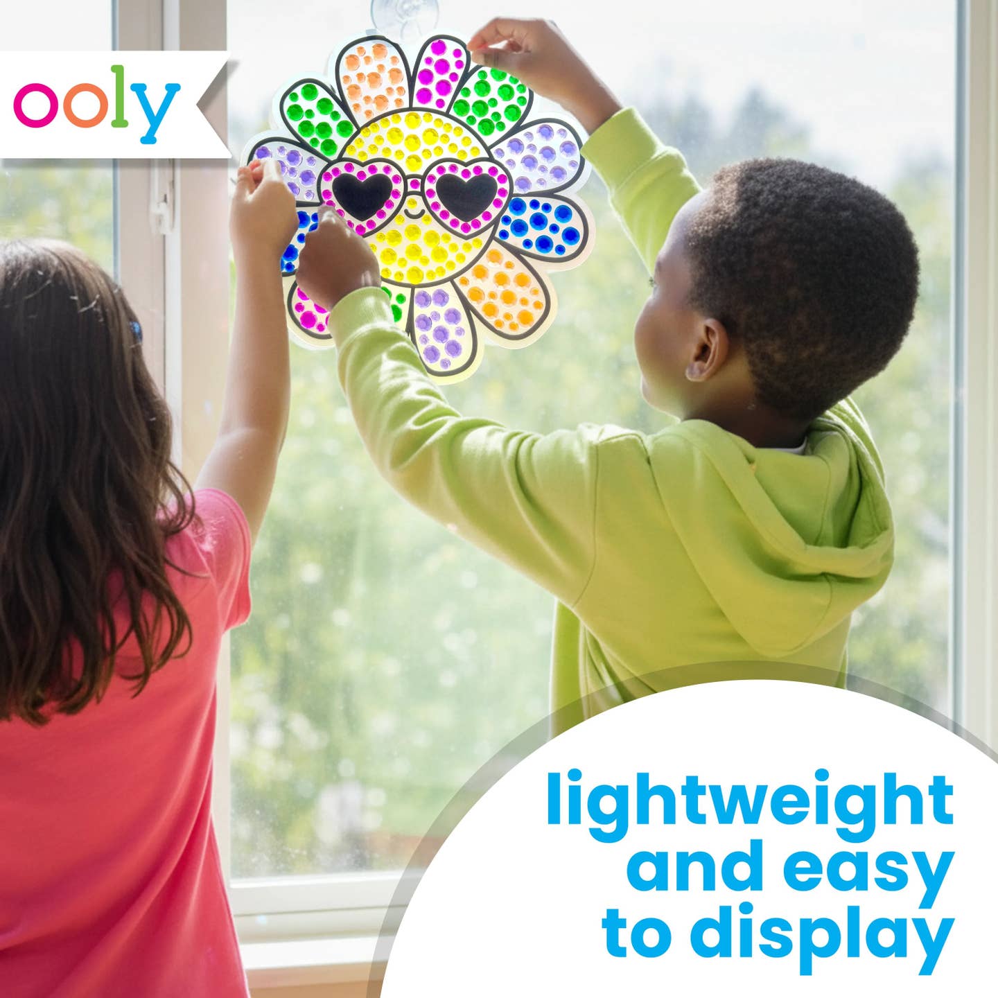 the back of two children, one with white skin and long brown hair and one with brown skin and short brown hair, hanging up the finished rainbow sun suncatcher in a brightly illuminated window
