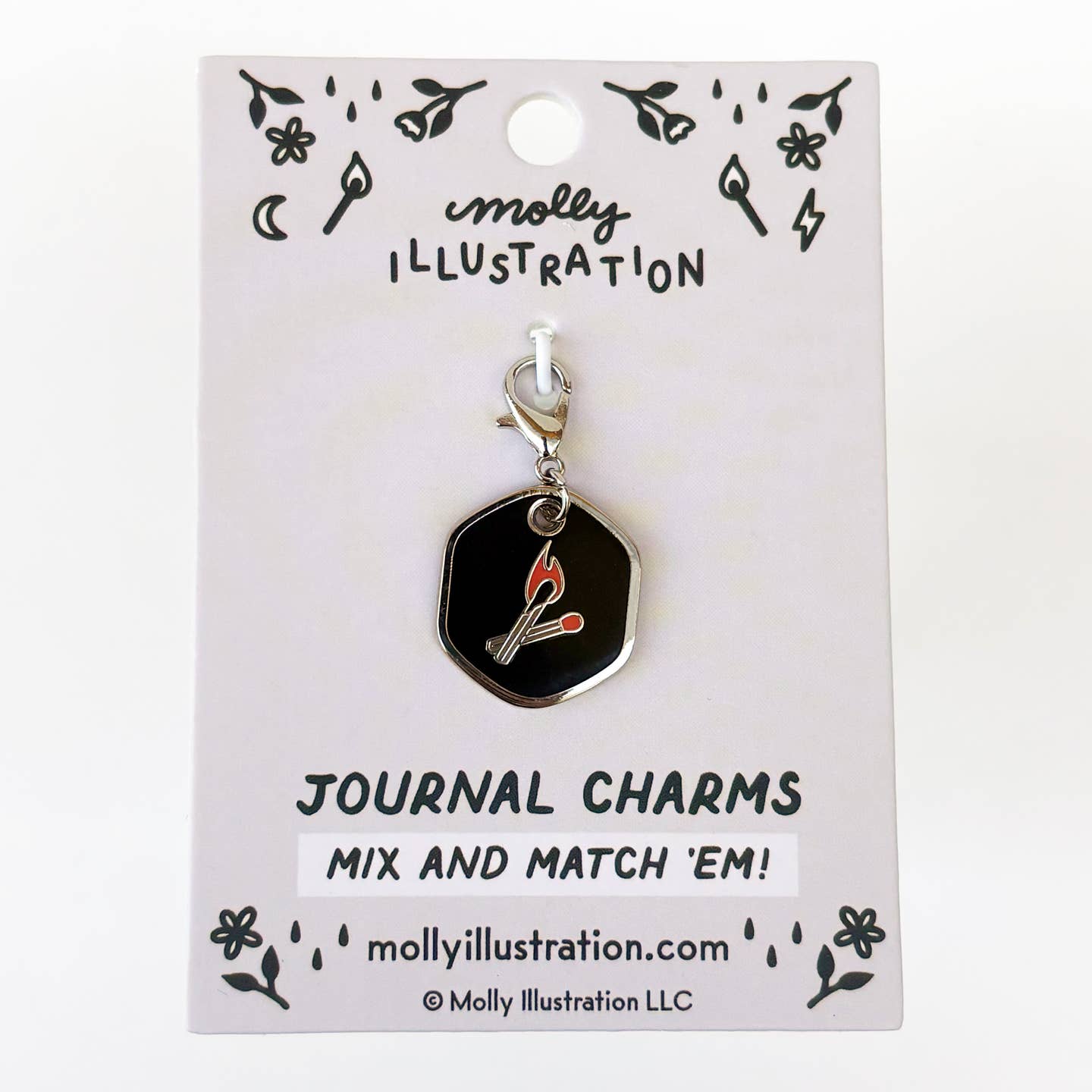 A black hexagonal journal charm with a silver clasp and a red-and-white match graphic, displayed on a white backing card labeled Molly Illustration.