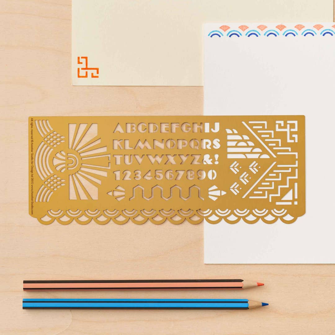 Decorative brass Art Deco ruler with letter and number designs on a wooden surface with a pencil and paper.