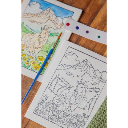 one painted and one unpainted version of the Glacier Mountain Goat watercolor card kit, with a paintbrush and dot card on a wooden background