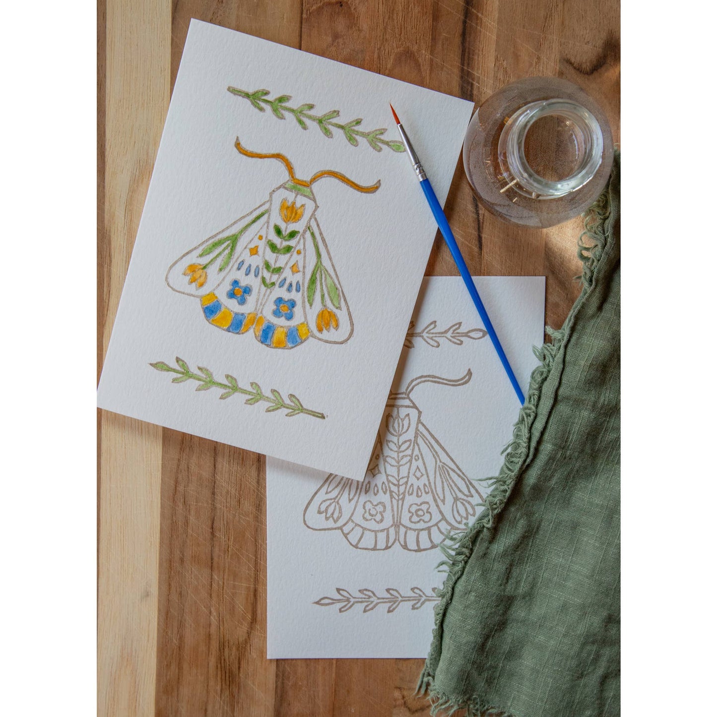 photo of one painted and one unpainted Folk Art Moth watercolor card kit with paintbrush and water glass on a wood background