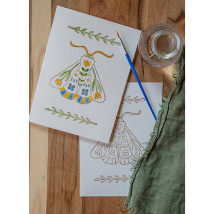 photo of one painted and one unpainted Folk Art Moth watercolor card kit with paintbrush and water glass on a wood background
