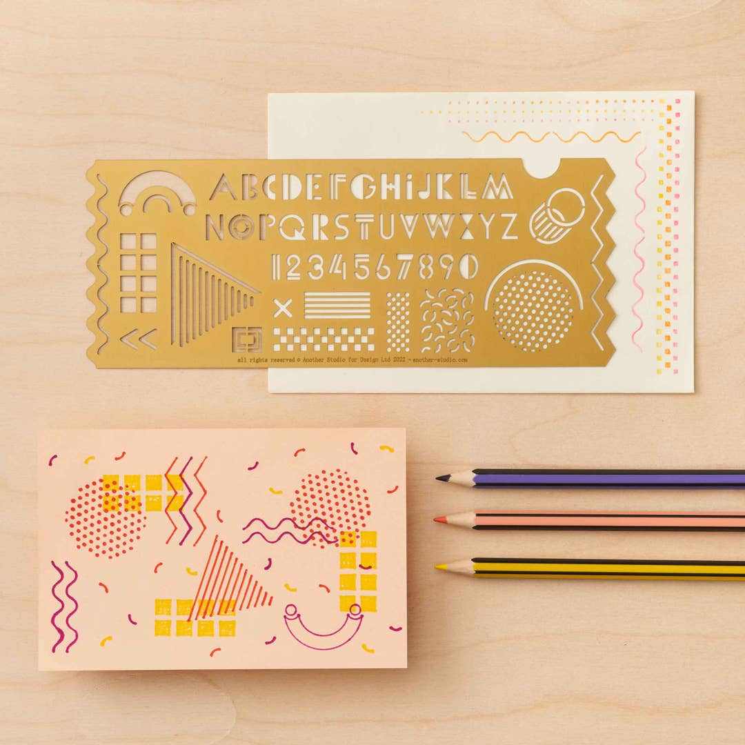 Decorative brass Memphis ruler with letter and number designs on a peach surface with a pencil and paper.