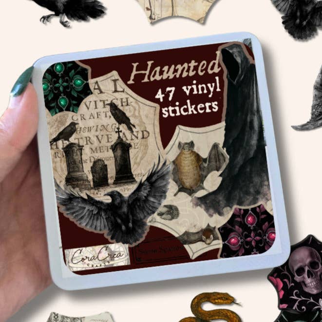 A hand holds a square tin of Coral Crea Crafts 'Haunted' vinyl stickers, showing a gothic collage design with ravens, tombstones, a cat, and the text 'Haunted 47 vinyl stickers'.