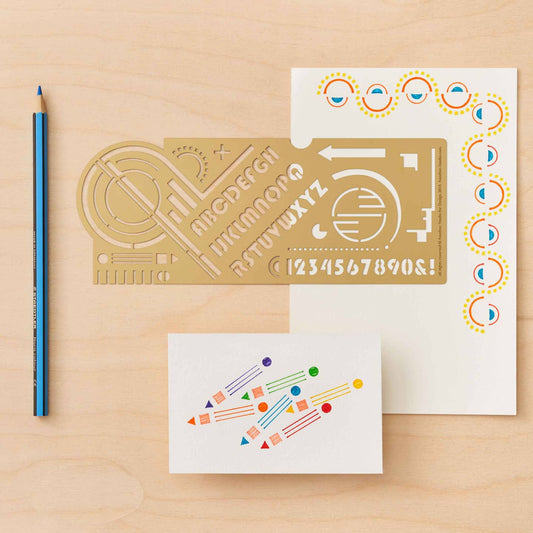 Decorative brass Bauhaus ruler with letter and number designs on a wooden surface with a pencil and paper.