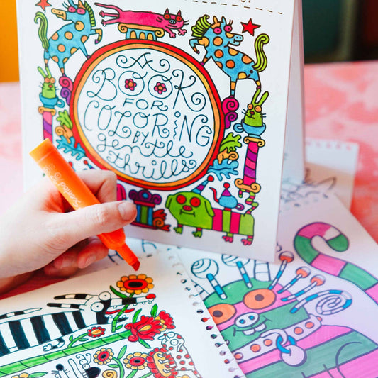 A colorful coloring book with whimsical illustrated characters and the title 'Book for Coloring by Gentle Thrills' stands open, with a white hand holding an orange marker over several coloring pages spread out on a pink surface.