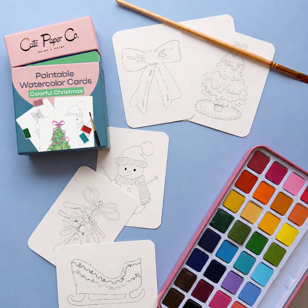 A flat-lay of a Christmas-themed paintable watercolor card set by Cute Paper Co. The set includes a pink and teal box, several white cards with black line drawings of holiday motifs (a bow, a Christmas tree, a snowman, a sleigh), a wooden paintbrush, and an open watercolor paint palette on a light blue background.