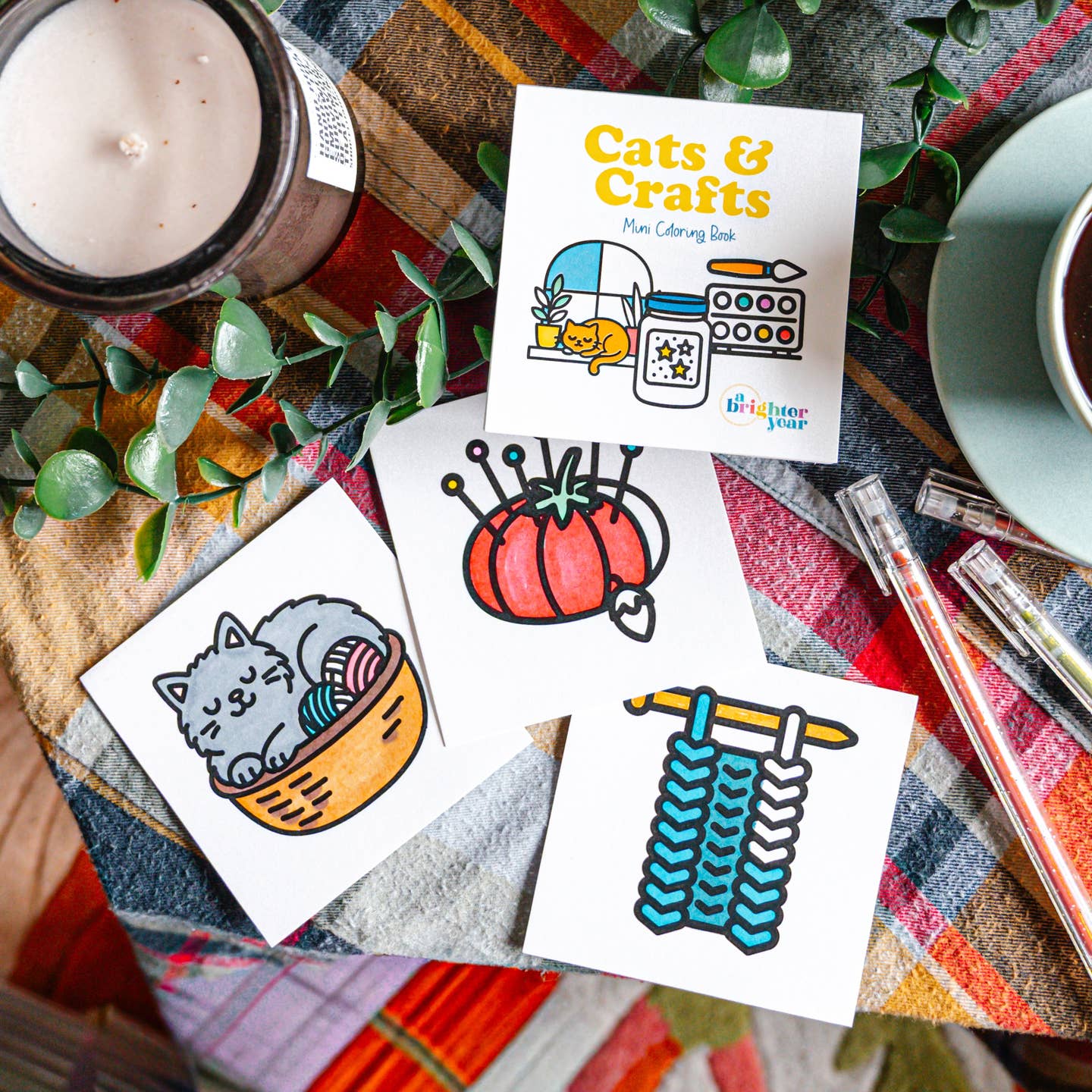 The Cats & Crafts coloring book  on a plaid tablecloth amongst a cup of tea, a candle, some gel pens, and completed coloring pages of a cat in a basket, a tomato pin cushion, and swatch of knitting