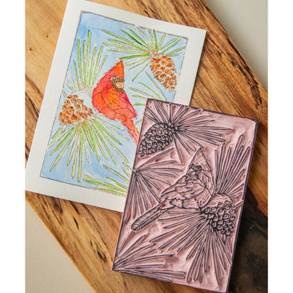A painted image of the Cardinal watercolor card kit laying alongside of the linoleum block on a natural wood background
