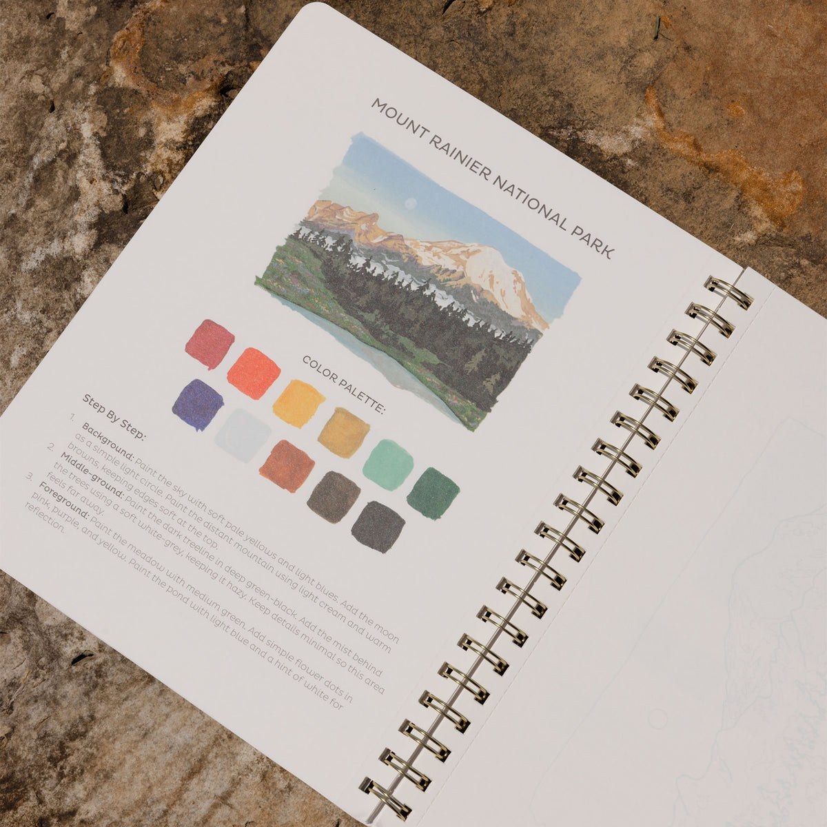 An open spiral-bound sketchbook lying on a textured stone surface, showing a printed page titled 'MOUNT RAINIER NATIONAL PARK' with a landscape illustration and a color palette section.