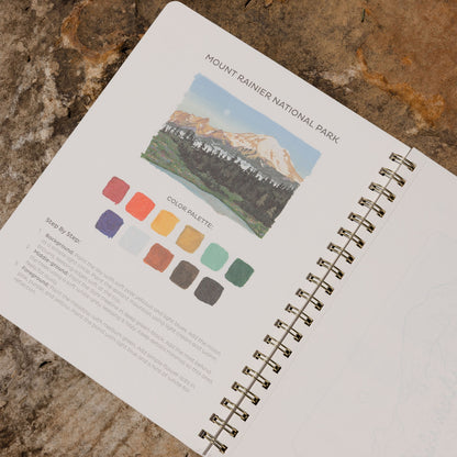 An open spiral-bound sketchbook lying on a textured stone surface, showing a printed page titled 'MOUNT RAINIER NATIONAL PARK' with a landscape illustration and a color palette section.