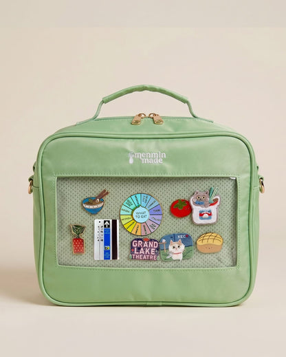 The Ultimate Journal Bag in  mint green lunch bag with a top handle, gold zipper, and a clear front window displaying various colorful enamel pins, set against a solid light beige background.