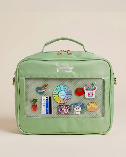 The Ultimate Journal Bag in  mint green lunch bag with a top handle, gold zipper, and a clear front window displaying various colorful enamel pins, set against a solid light beige background.