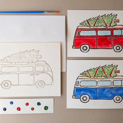 one unpainted and two painted versions of the Van watercolor card kit, one van is painted red, while the other is painted blue, each with a christmas tree tied to the top. A paintbrush and dot card lays on top.