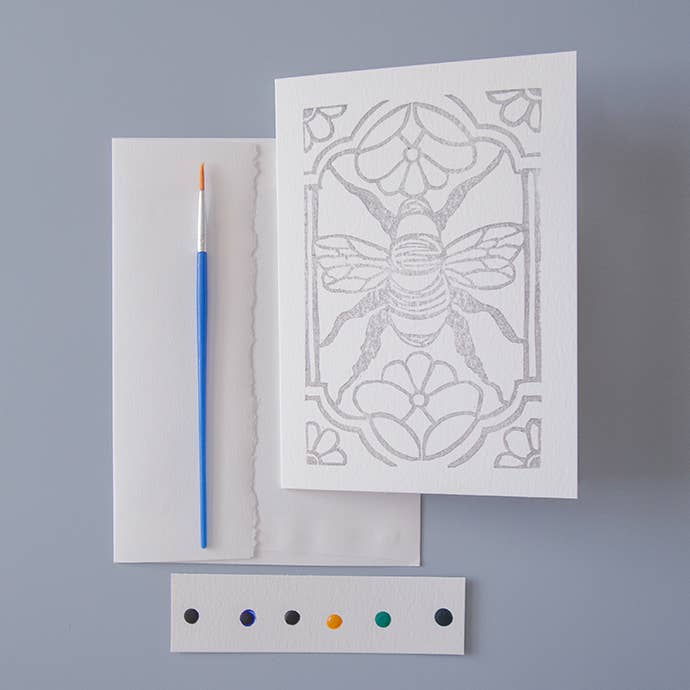 image of a paintbrush, watercolor dot card, and unpainted linocut artwork outline of a stylized bee with flowers