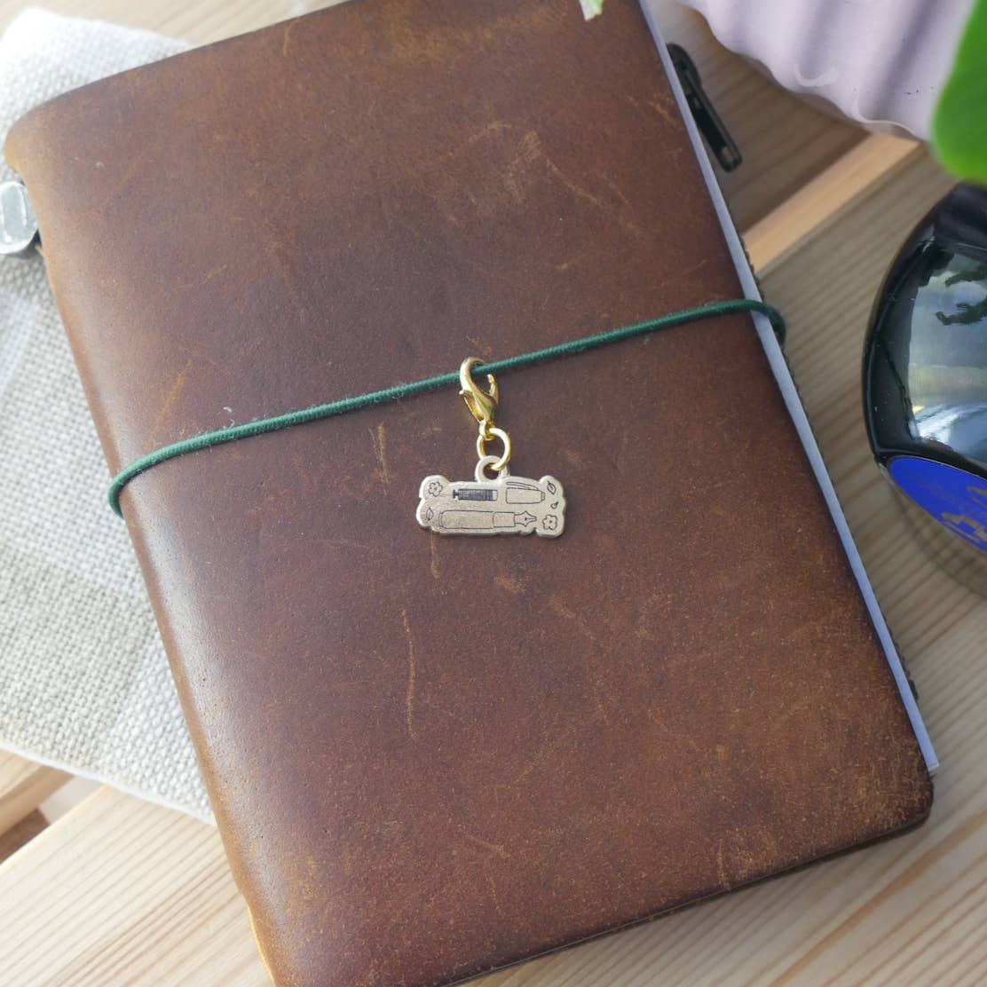Small brass "Handwritten with Love" charm engraved with an illustration of a fountain and parts with a gold-tone clasp attached to a brown leather Traveler's style jounral.