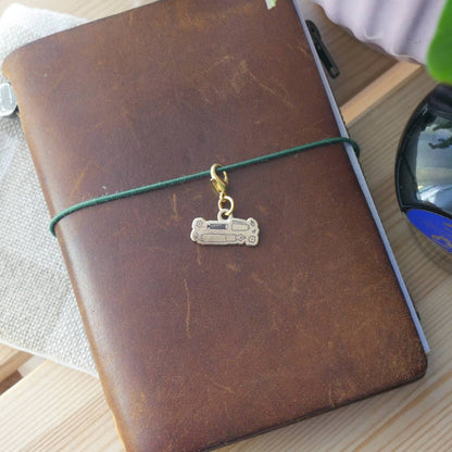 Small brass "Handwritten with Love" charm engraved with an illustration of a fountain and parts with a gold-tone clasp attached to a brown leather Traveler's style jounral.