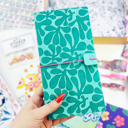 A hand with red nail polish holds a teal and green patterned traveler notebook or planner cover with a pink elastic band closure. The background is a colorful, slightly blurred stationery display.