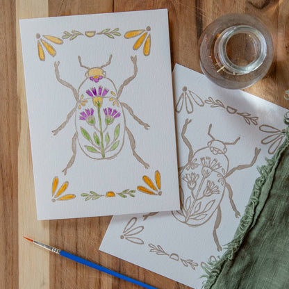 image of one unfinished and one finished example of a folk art beetle with purple and yellow flower details