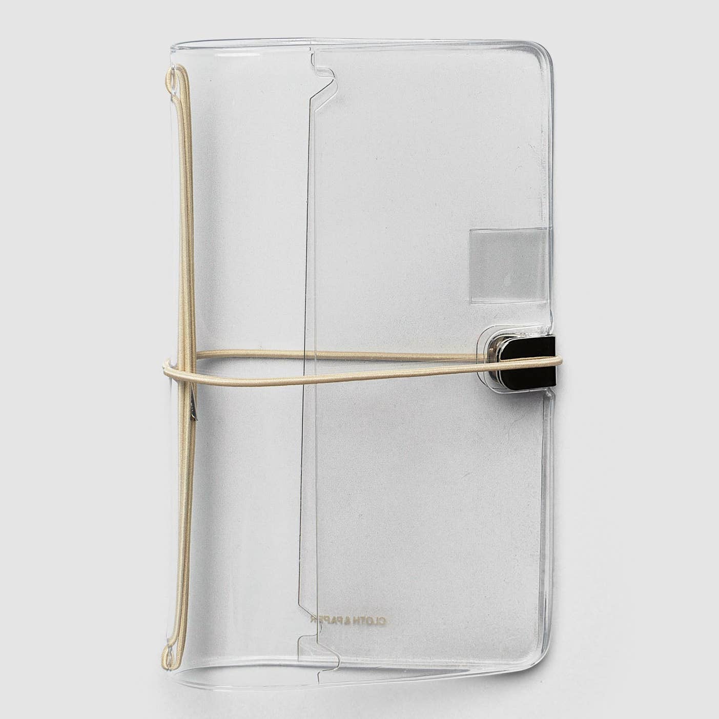 A clear, transparent notebook cover with a minimalist design, featuring a gold-colored elastic band closure and a black metal clasp, shown on a light gray background.