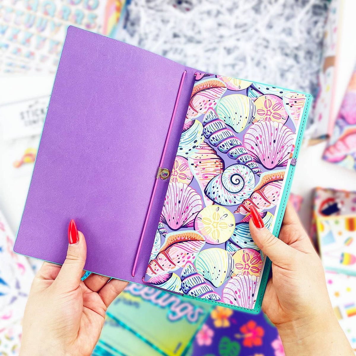 A person holds an open purple notebook or planner cover. The right page is decorated with a colorful seashell pattern in pastel shades of pink, purple, blue, and yellow.