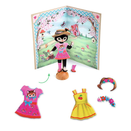 black cat figurine in front of a scenic background card wearing a pink dress with a kitten on it, pink wellies, and a straw hat. At the cat's feet is another yellow dress and floral crown that can be put on alternatively.