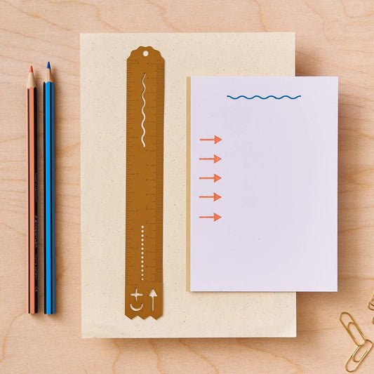 A flat-lay view of the brass straight edged ruler with measurement markings, a scalloped line stencil, a dot line stencil, and arrow, plus, and crescent stencil, placed next to a white notepad with printed arrows and squiggles, on a beige paper sheet over a light wood background. Two colored pencils and gold paperclips are visible on the side