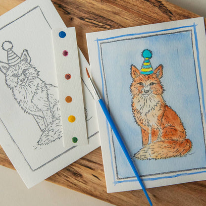 Card with an illustration of a fox wearing a party hat and watercolors and paintbrush on a wooden surface