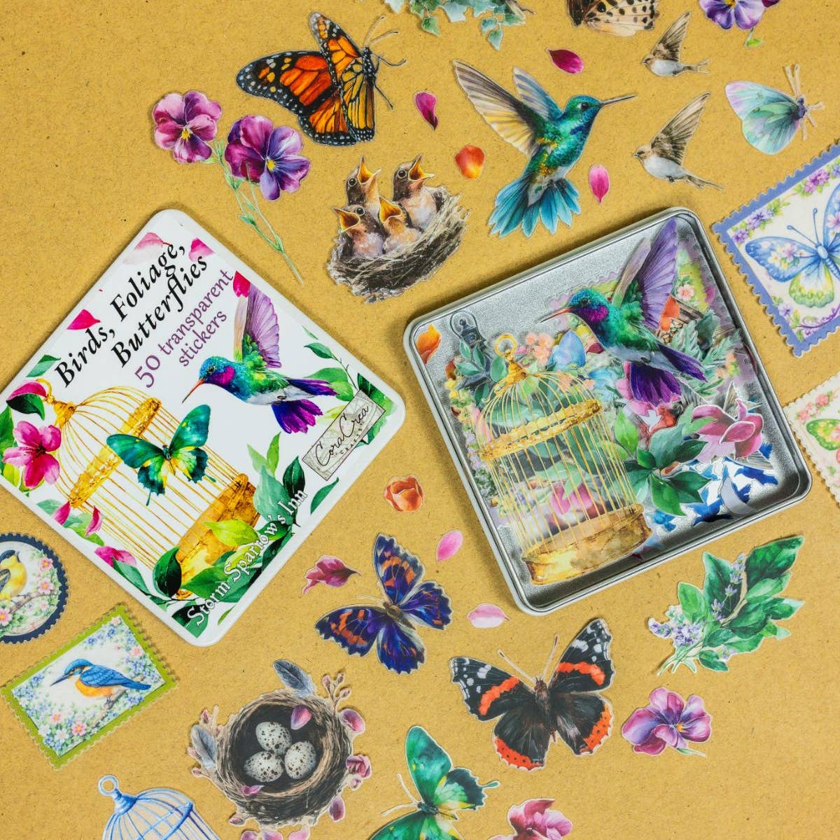A styled flat-lay of bird and butterfly-themed stickers on a tan textured background. Two metal tin boxes are visible, one labeled 'Birds, Eggs, Butterflies 50 temporary stickers,' surrounded by scattered die-cut stickers including hummingbirds, butterflies, flowers, nests, and leaves.