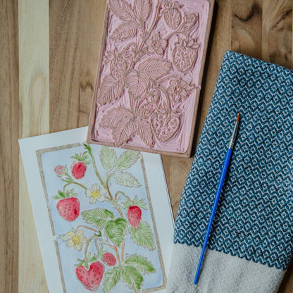 photo of the original linoblock and a completed example of the strawberries art, painted with red berries, white flowers on a blue background