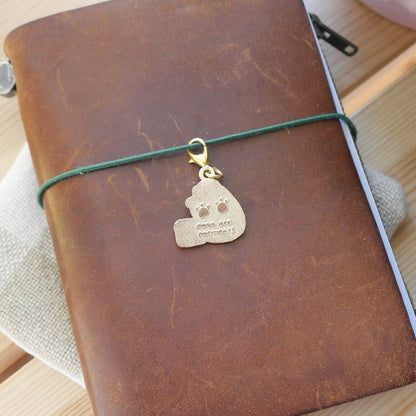 Back of the brass "Paw Prints" charm engraved with paw prints with a gold-tone clasp attached to a brown leather Traveler's style jounral.