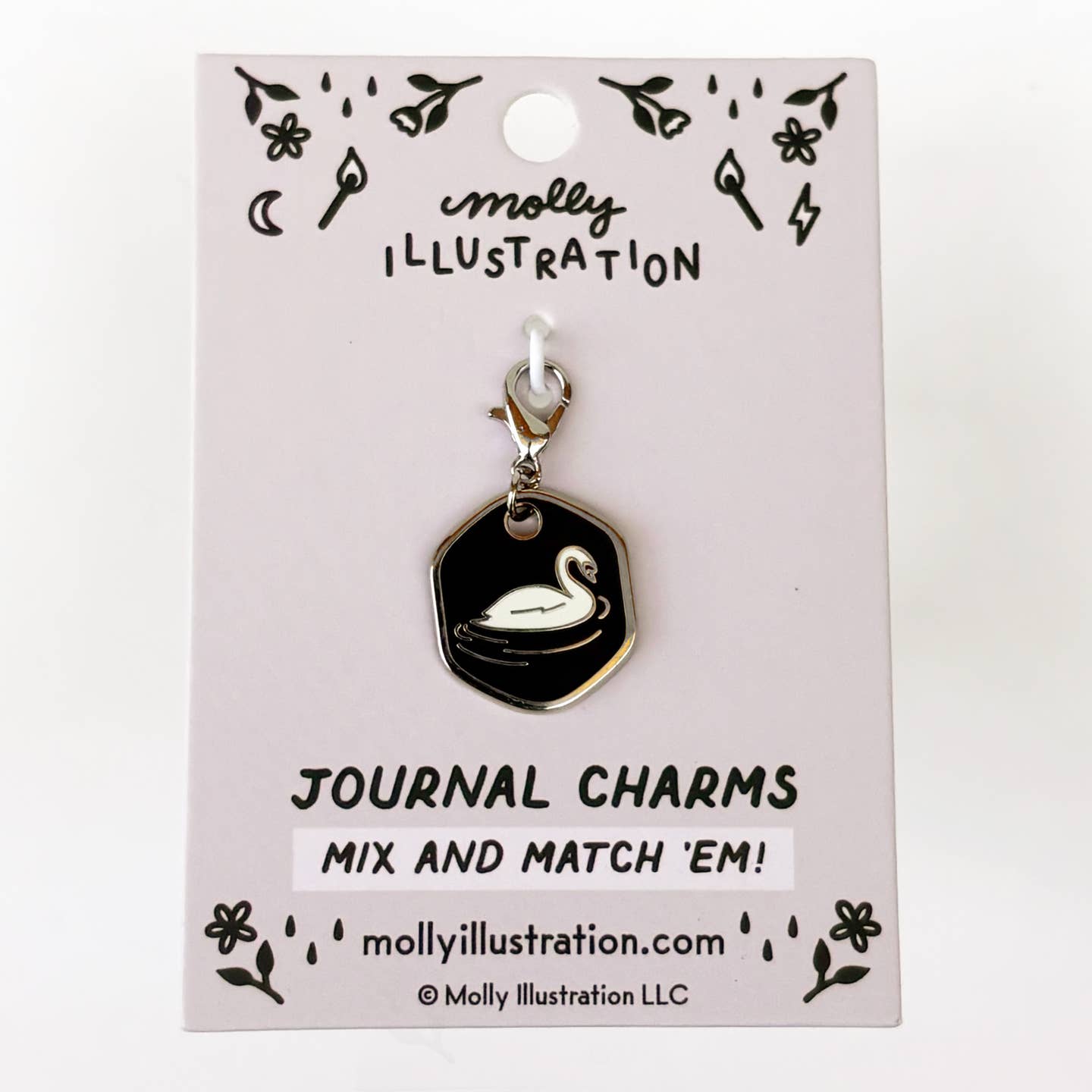 A journal charm by Molly Illustration, featuring a black hexagonal pendant with a white swan, attached to a silver clasp and displayed on its light pink packaging card.
