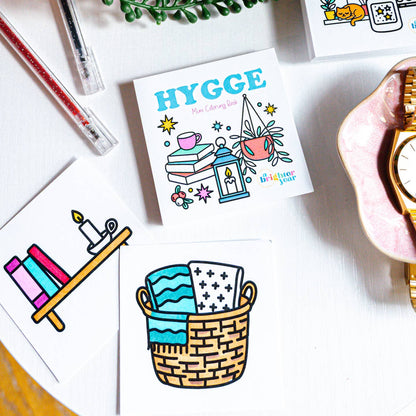 images from the interior of the Hygge coloring book featuring a basket full of blankets and a book shelf with books and a candle