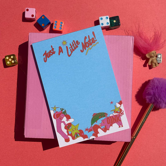 A flat-lay image of a pastel blue notepad with the text 'Just A Little Note!' and whimsical pink cartoon characters like a whacky waver and a pig at the bottom, on a red and pink background. The scene is styled with colorful dice, a purple pom-pom pen, and a small troll doll on a solid pink background.
