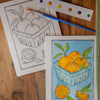 Card with an illustration of oranges in a produce box with watercolors and a paintbrush on a wooden surface