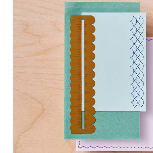 A scalloped edged brass ruler with metric and imperial markings laid over a white sheet with blue fish-scale patterned edges, placed on a teal cardstock sheet over a light wood surface.
