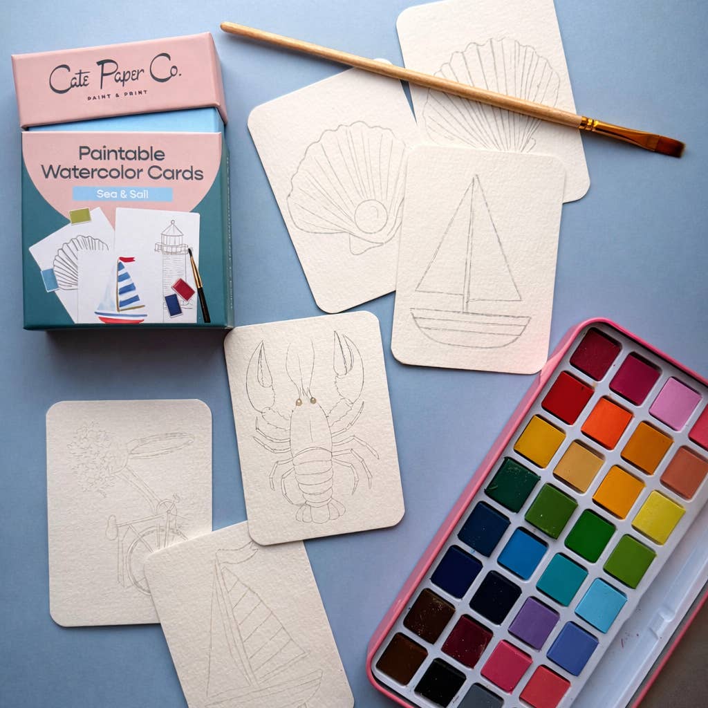 A flat-lay of a watercolor card painting set by Cafe Paper Co. on a light blue background, featuring cream-colored watercolor cards with shell and sailboat sketches, a pink watercolor paint palette, a wooden paintbrush, and the product box.